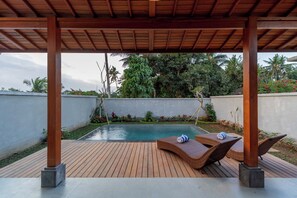 A covered outdoor area with a pool, lounge chairs, and a wooden deck. - Sila Living Ubud (Sukawati)