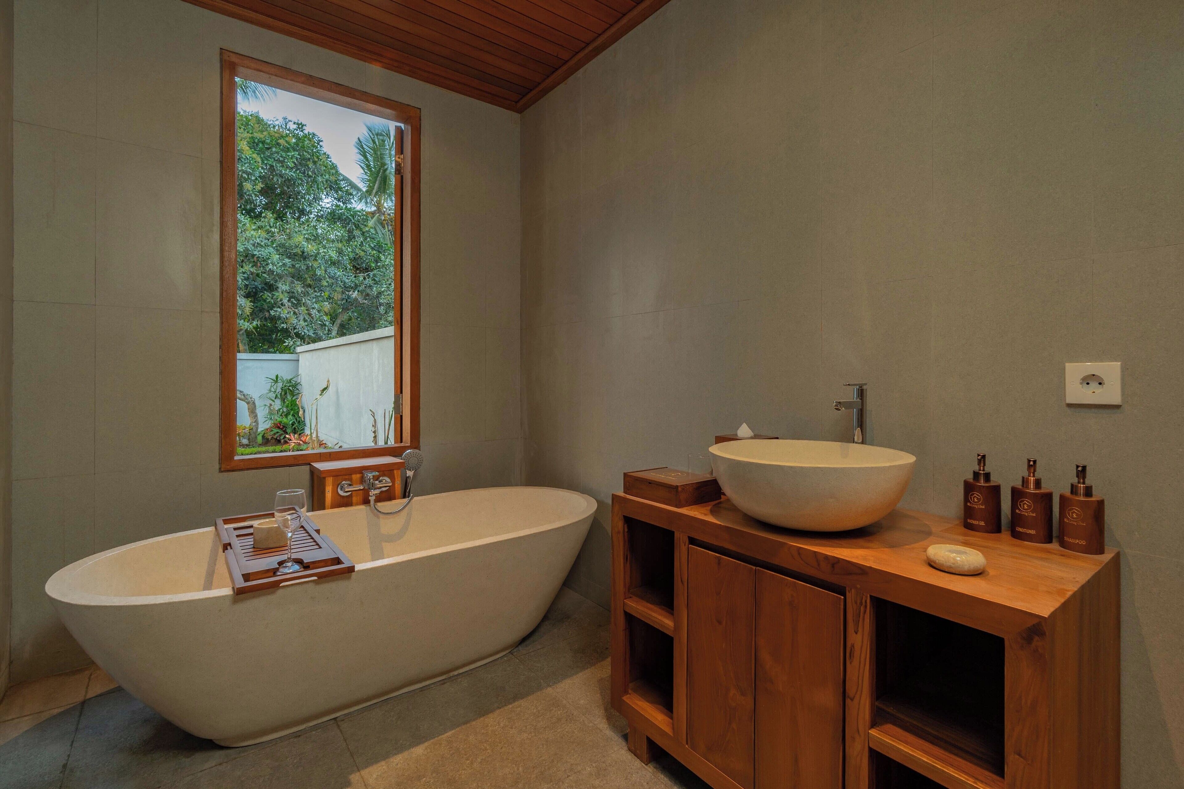 A modern bathroom with a freestanding bathtub, a wooden vanity, and a window providing a view of greenery.