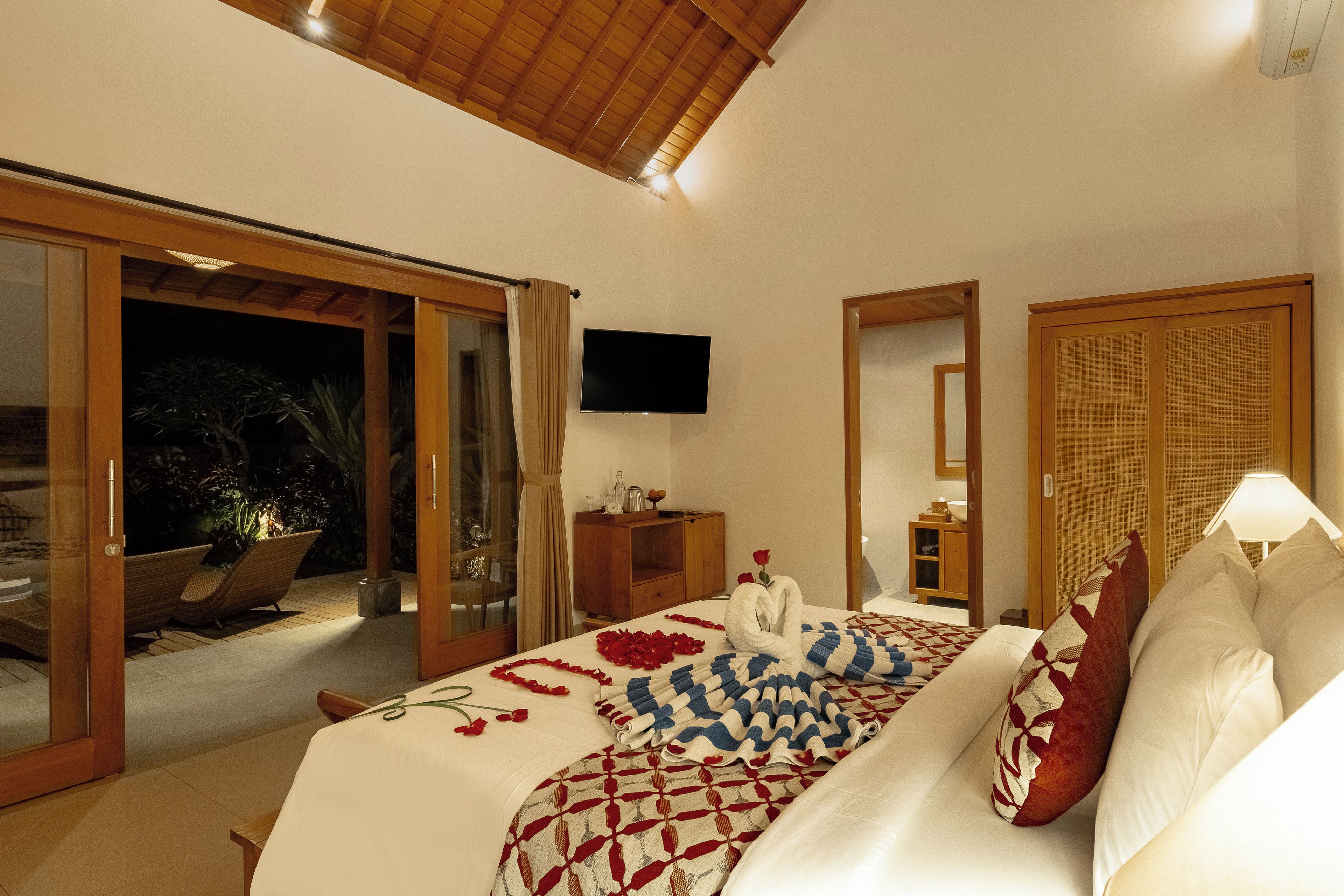 A bedroom with a bed, a television, a wooden wardrobe, and a sliding glass door leading to an outdoor area.