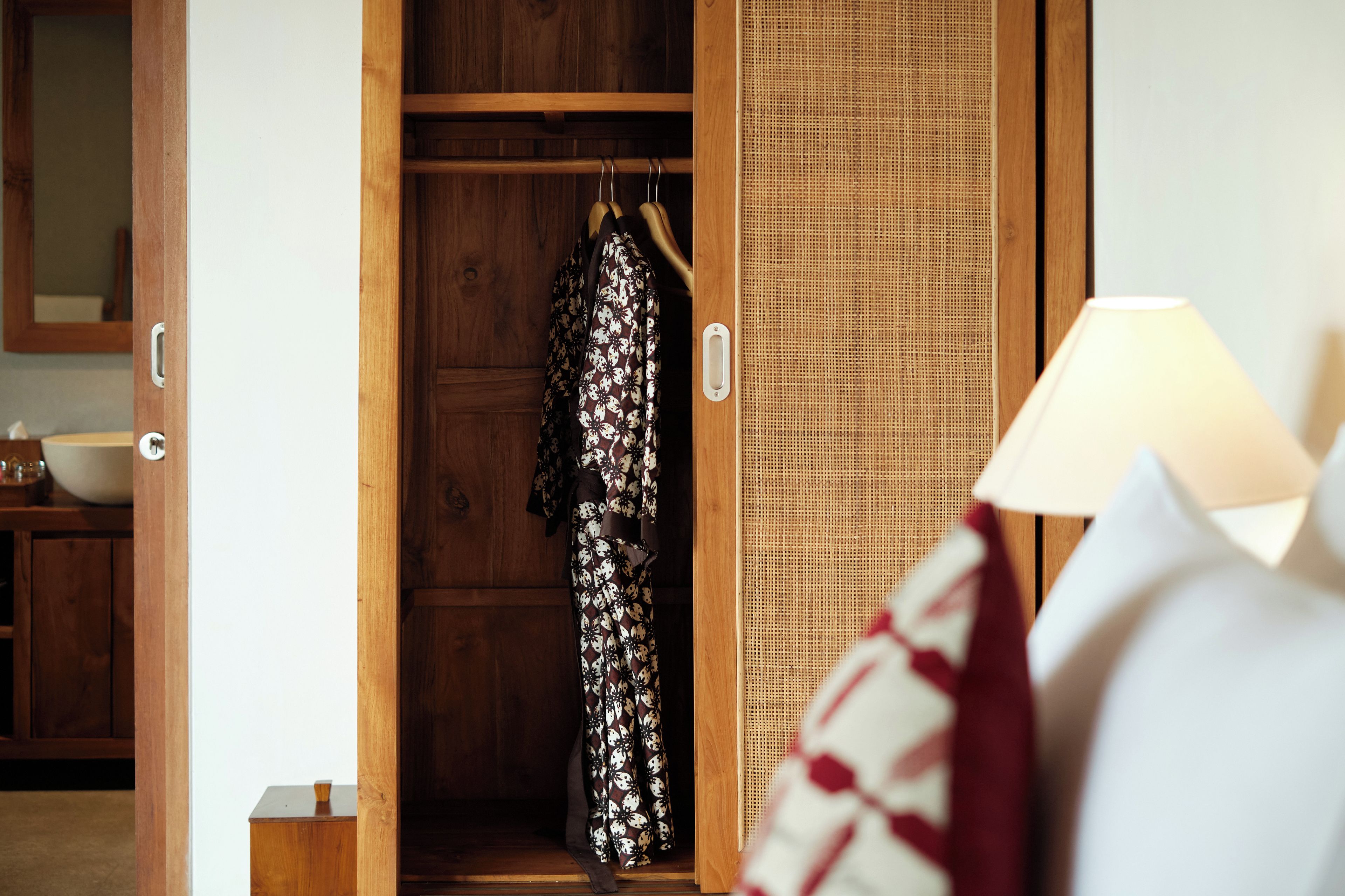A wooden wardrobe with a hanging garment, a wicker basket, and a bedside lamp.