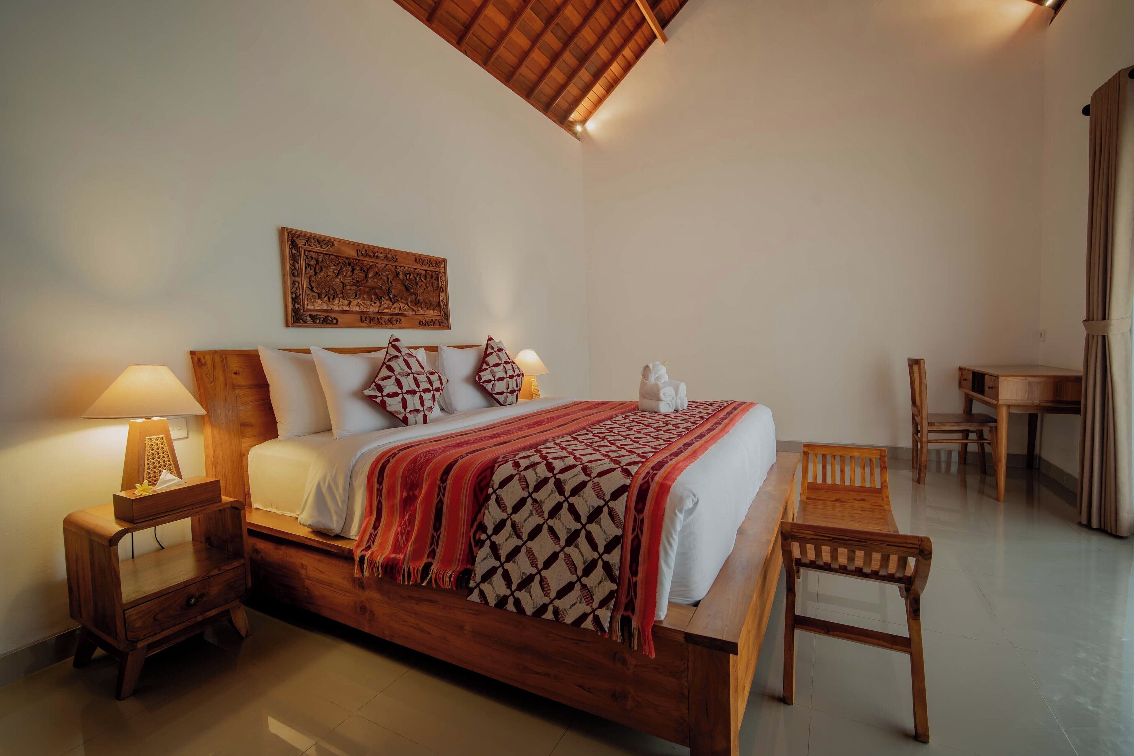 Een houten bed met een bedsprei met patroon, een nachtkastje met een lamp en een houten stoel.