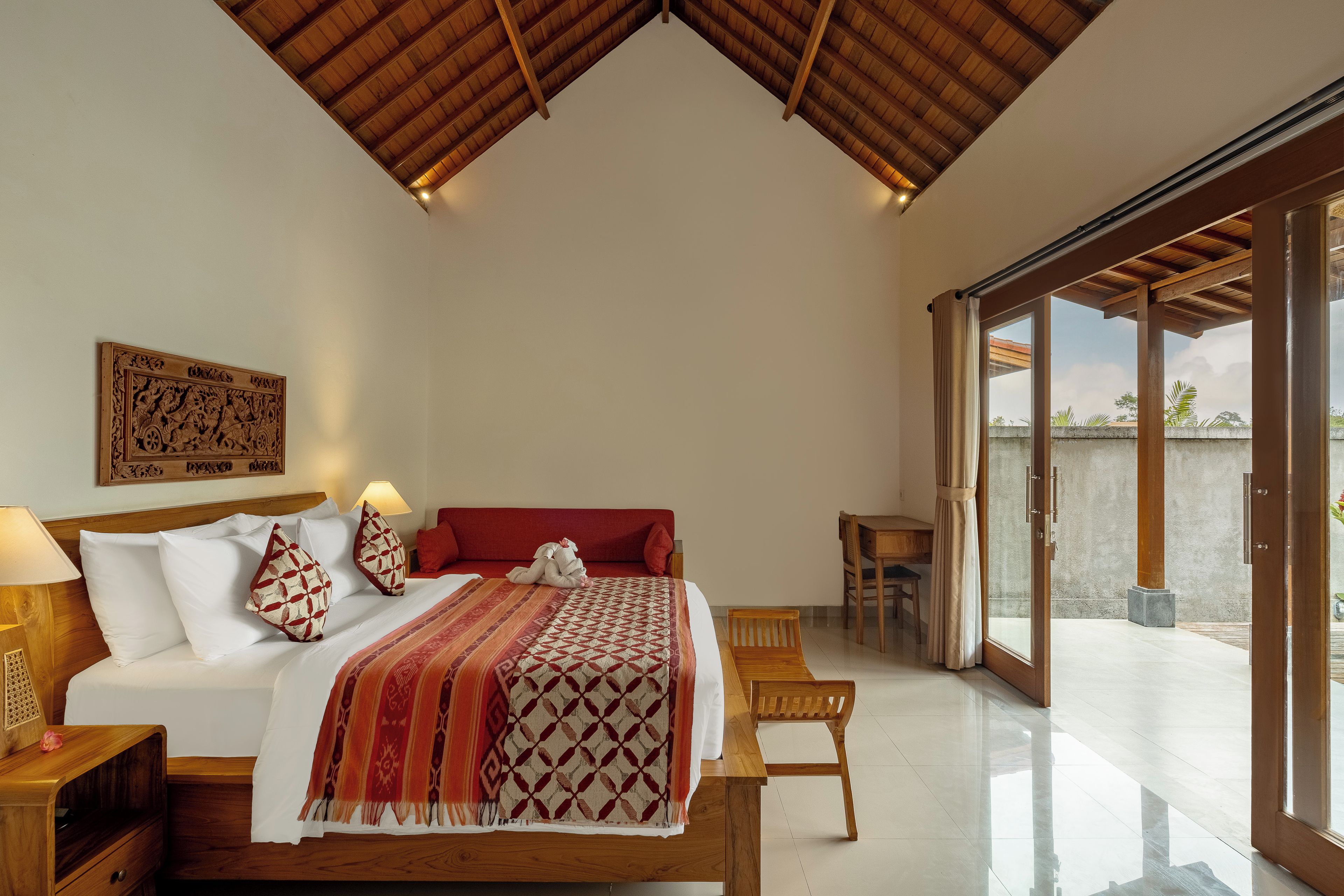 A bedroom with a large bed, a wooden headboard, a nightstand with a lamp, a red sofa, a wooden chair, and a view through a glass door.
