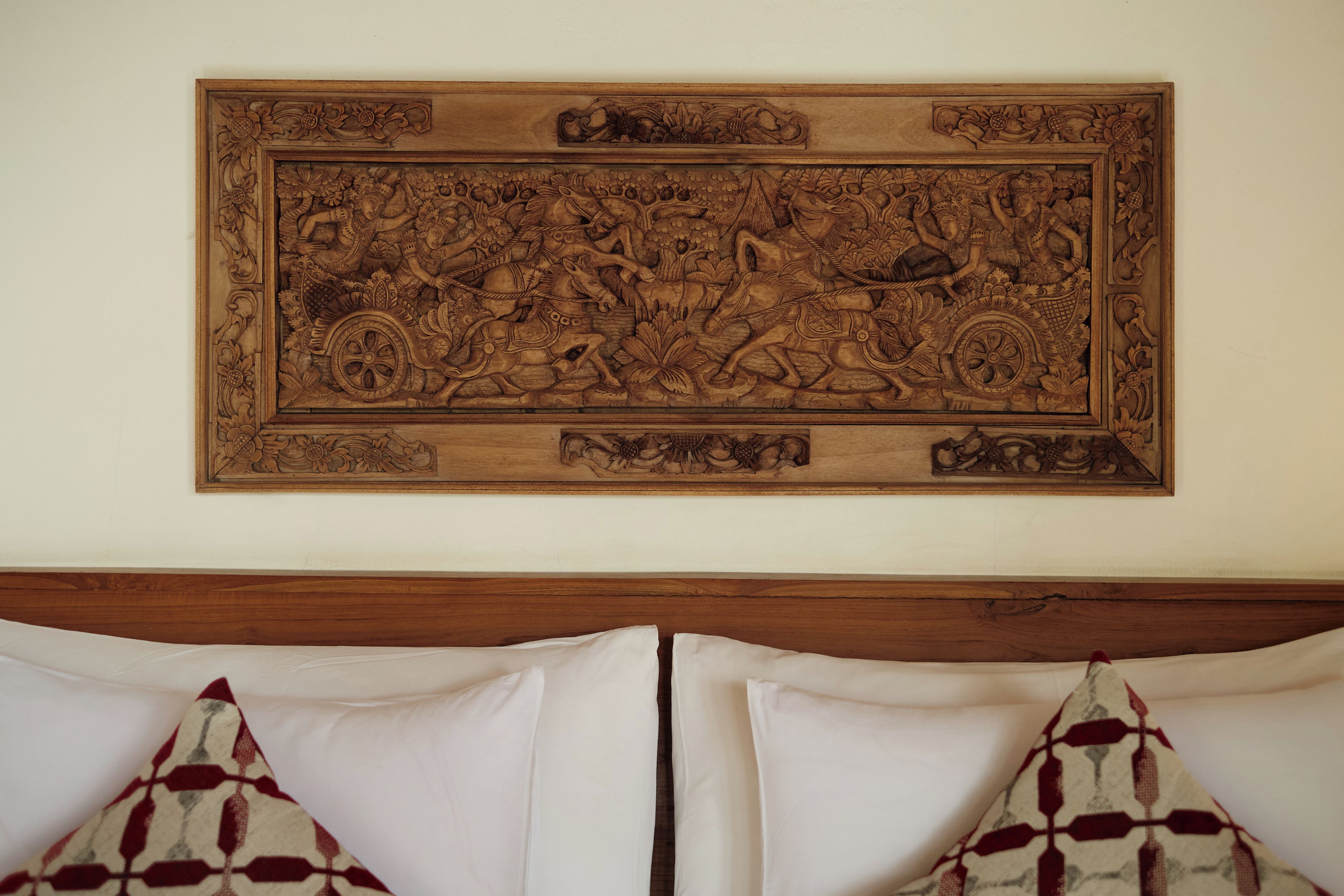 A carved wooden panel with intricate designs, mounted on a wall above a bed with white bedding and patterned pillows.