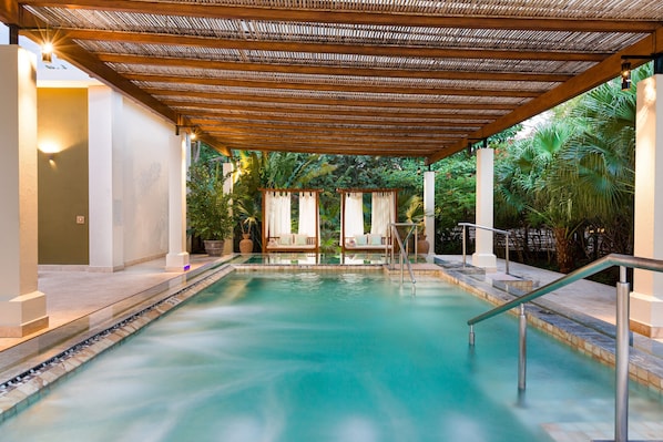 A serene indoor pool area with a wooden pergola, lounge chairs, and a glass barrier.