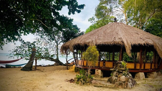 A thatched-roof hut with a wooden deck, surrounded by trees and rocks, overlooking a beach.