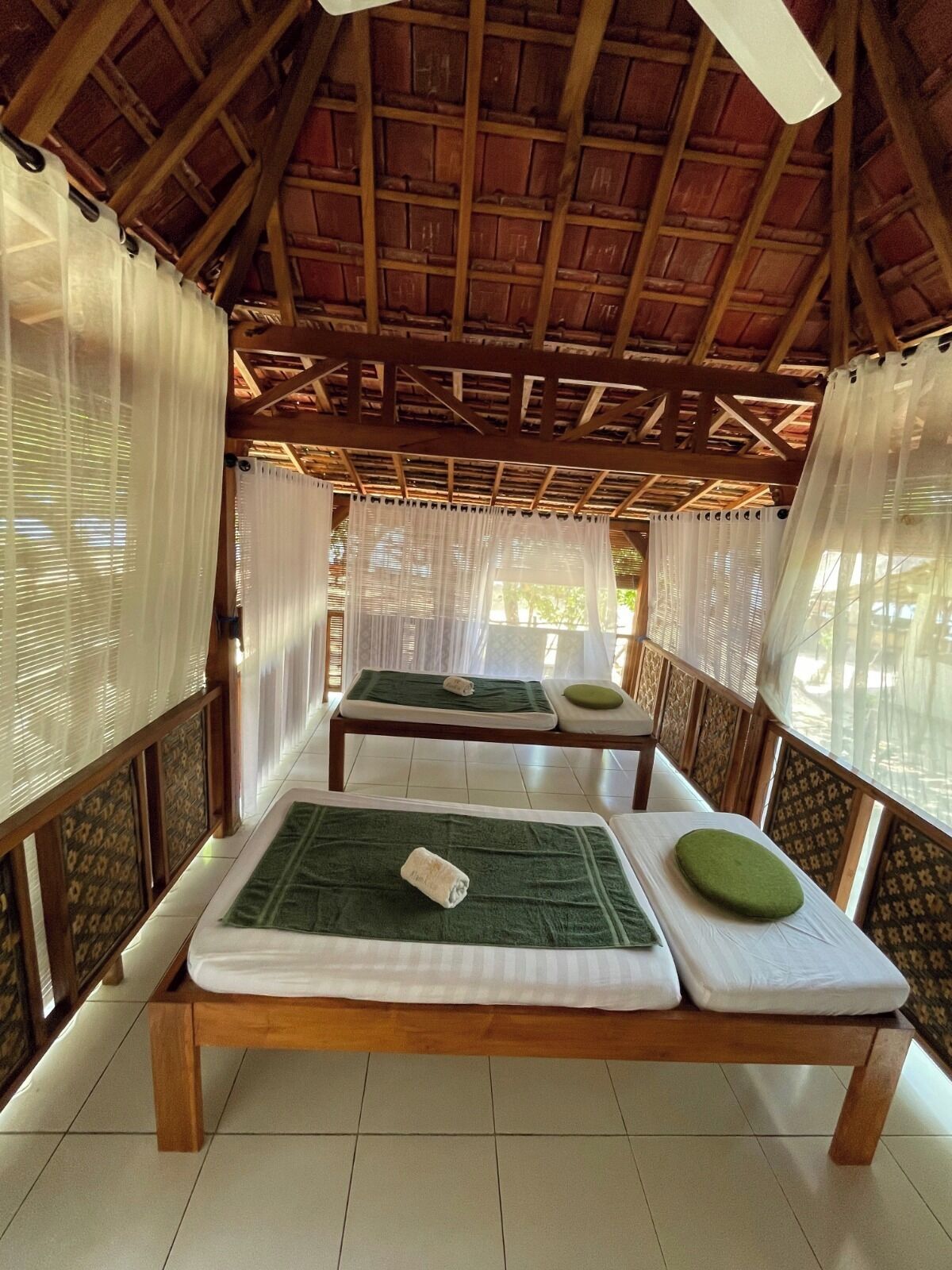 Ein Zimmer mit Holzbalken und einem Reetdach, ausgestattet mit zwei Massagetischen, grünen Matten und weißen Handtüchern.