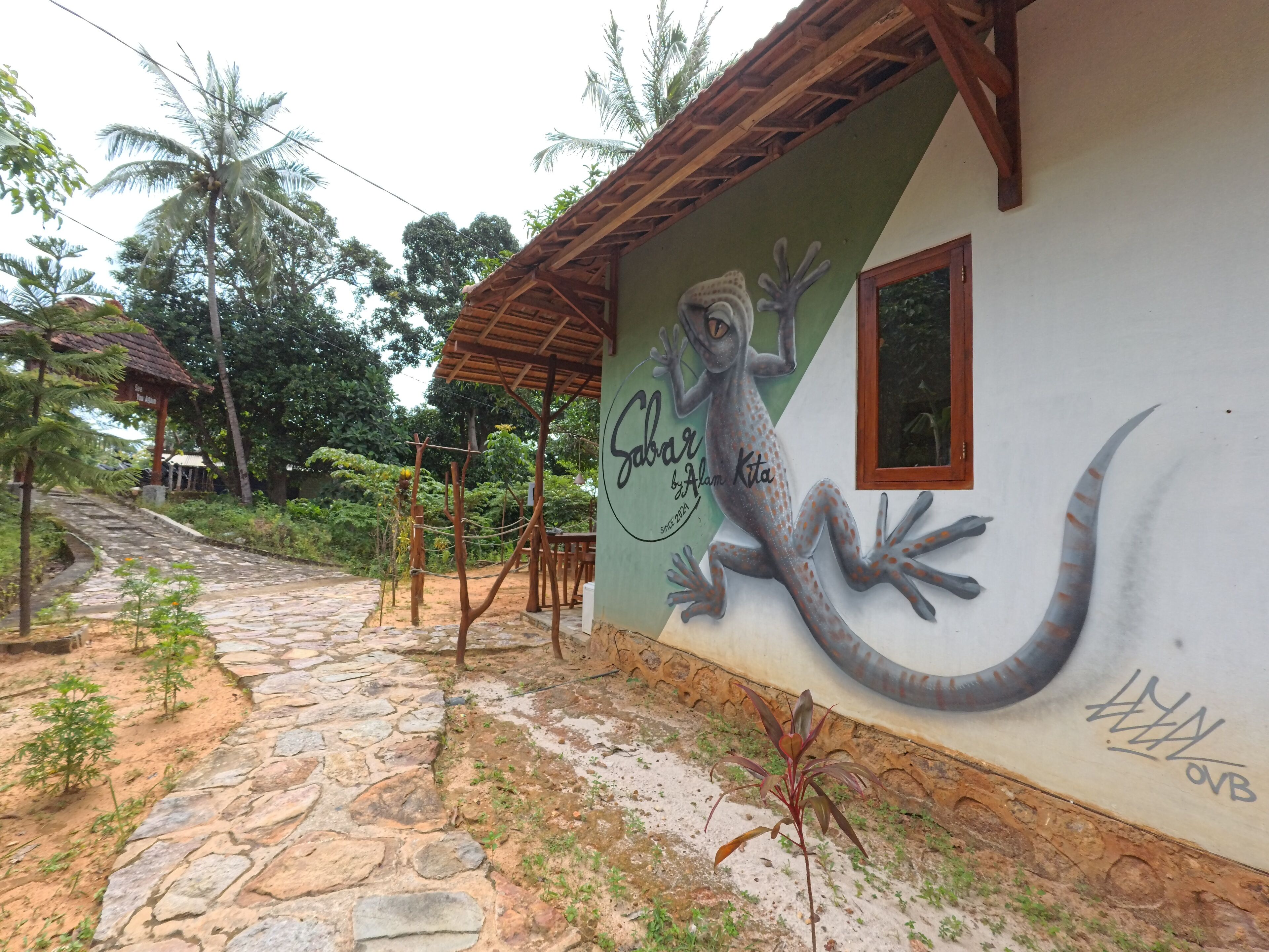 Un bâtiment rustique orné d’une fresque représentant un gecko, un sentier en pierre et une structure en bois en arrière-plan.