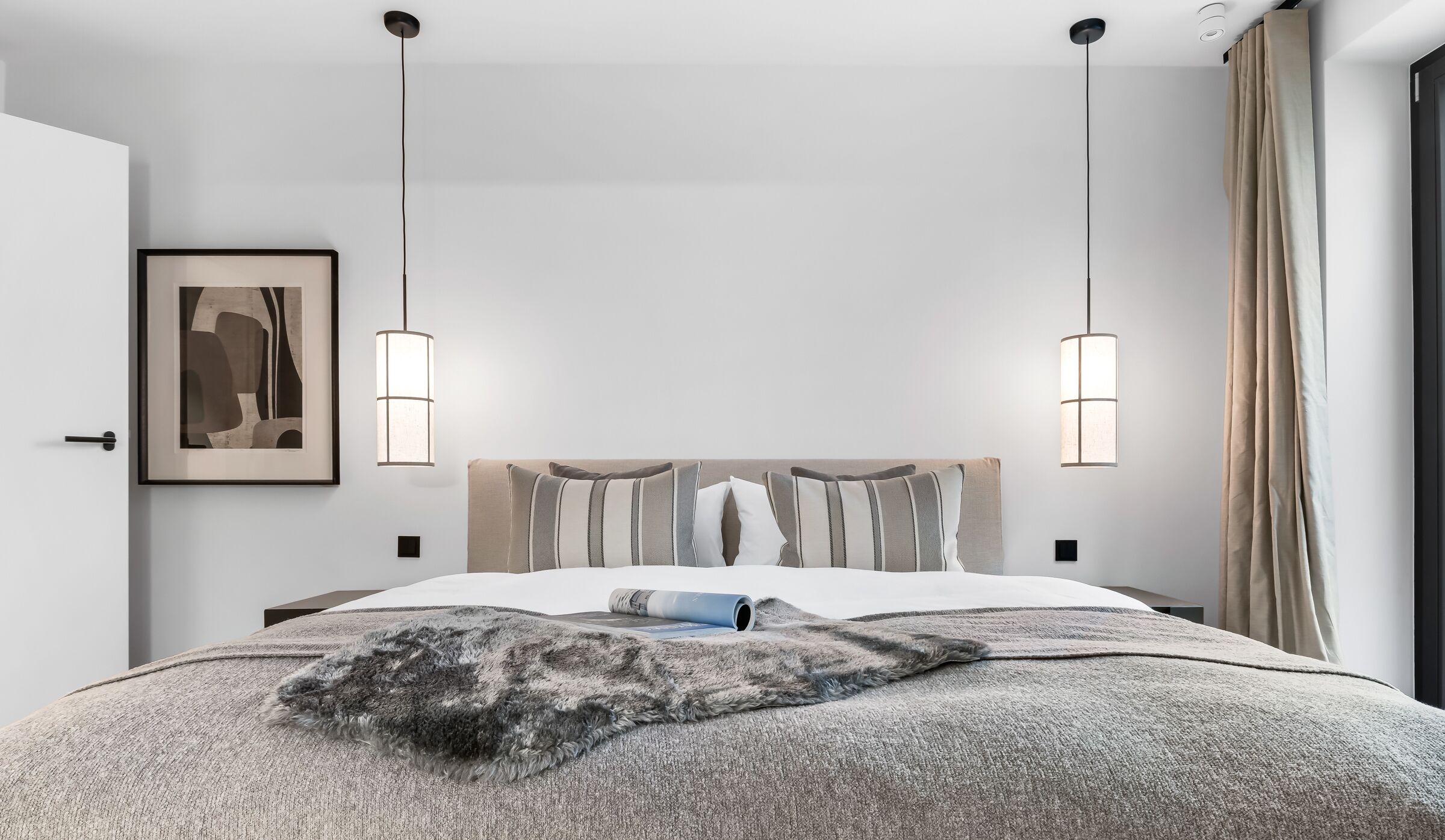 Quarto moderno com cama grande, duas luminárias pendentes, uma obra de arte emoldurada e vista para o exterior.