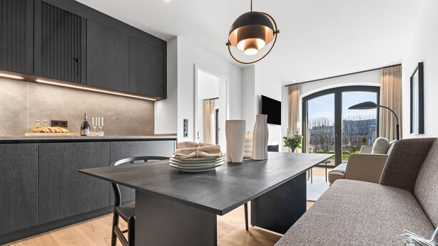 A modern kitchen with dark cabinetry, a central island, and a dining area with a sofa and table. - Manhattan Suit – New Luxury Spread Across 2 Floors with Finnish & Infrared Sauna (List auf Sylt)