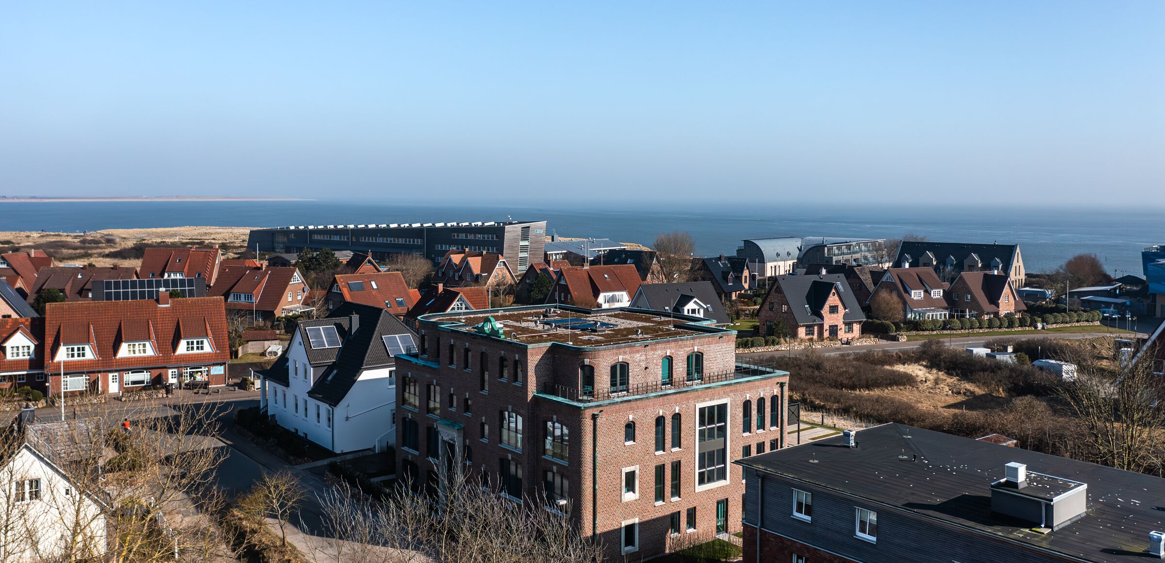 A residential area with houses, a large building with a flat roof, and a view of the sea.