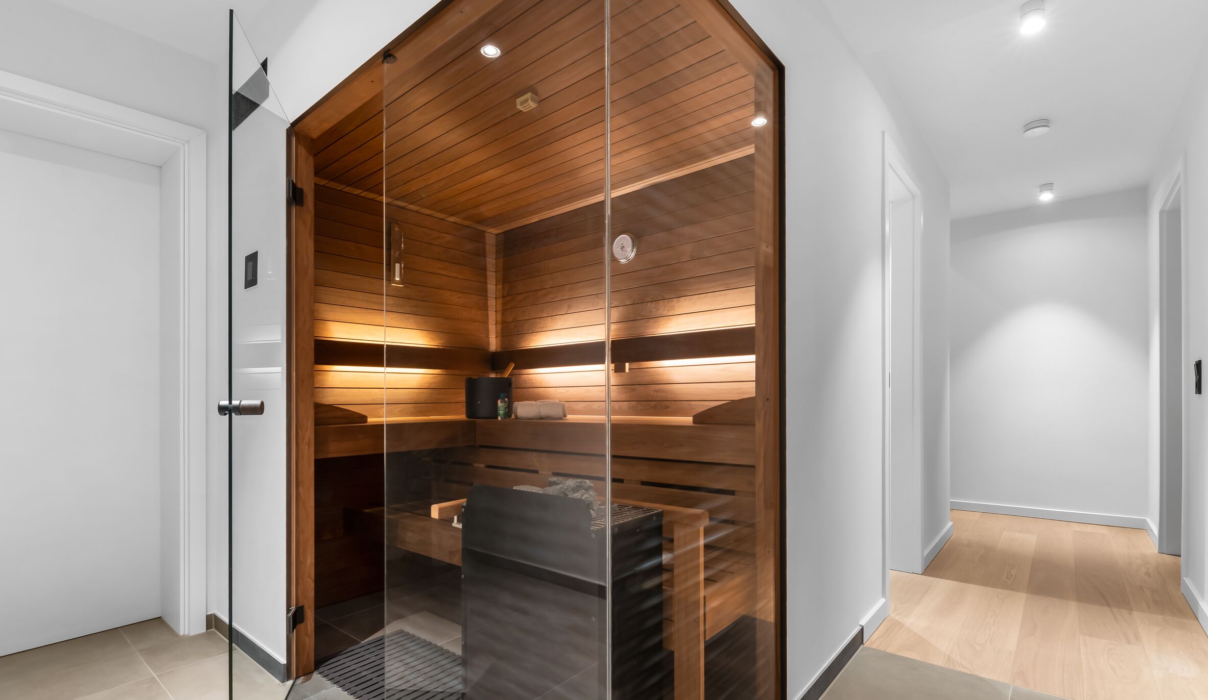 A modern, well-lit sauna with wooden slats and a glass door, located in a hallway with light-colored walls and wooden flooring.