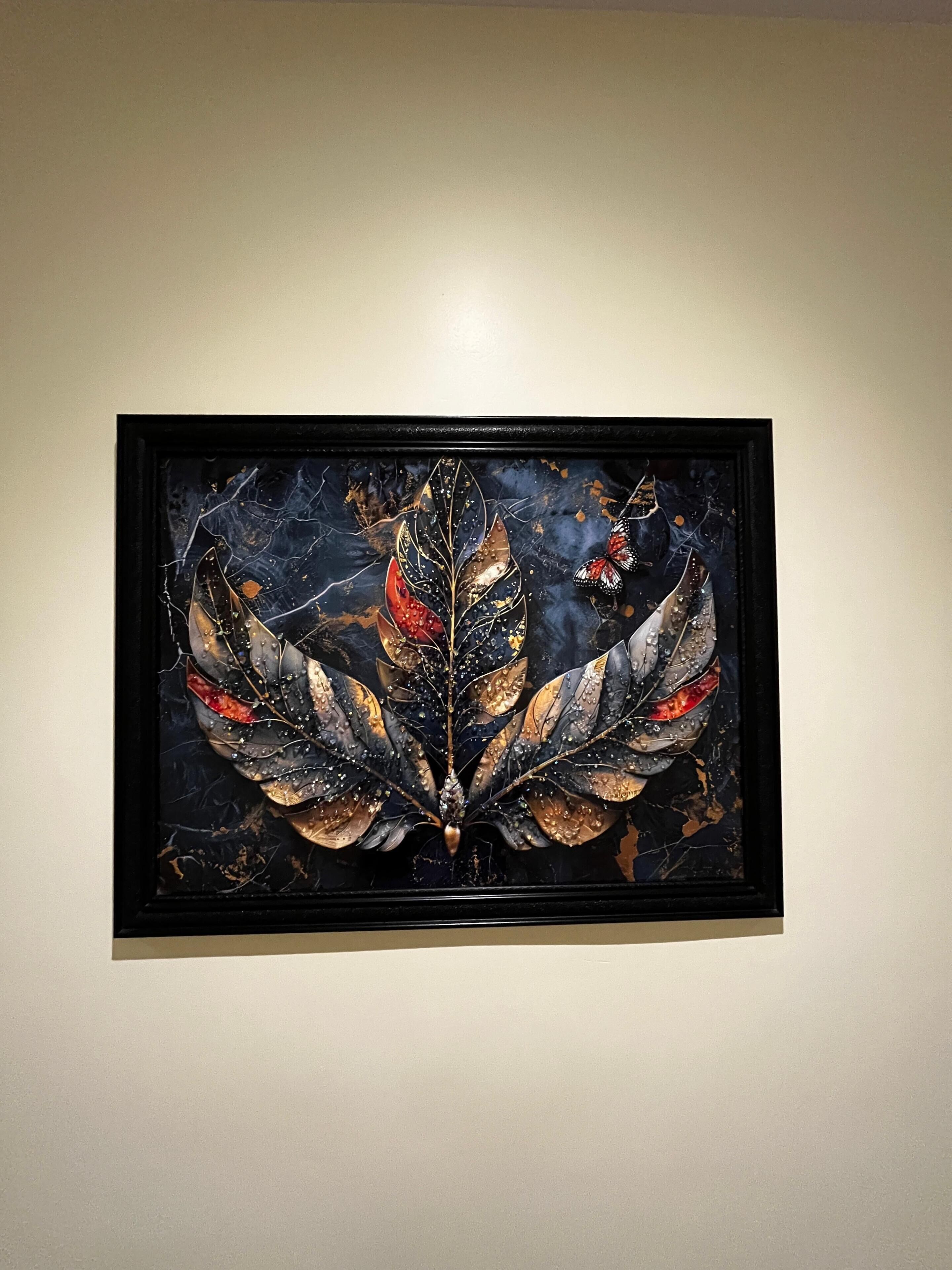 A framed artwork with a central leaf motif and butterfly elements, displayed on a plain wall.
