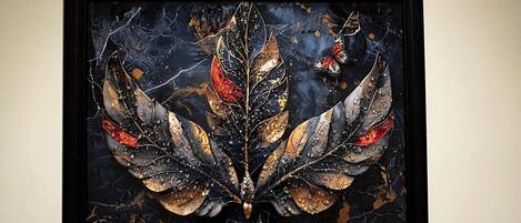 A framed artwork with a central leaf motif and butterfly elements, displayed on a plain wall.