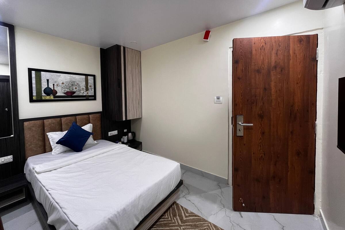 A hotel room with a bed, a wooden door, a nightstand, a framed picture on the wall, and a small table.