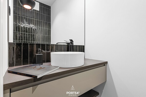 A modern bathroom with a white sink, dark tiles, and a magazine on the counter. - East Village Suite luxurious apartment with an fireplace and infrared shower. (List auf Sylt)