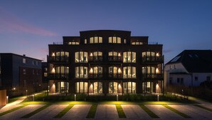 Un immeuble à plusieurs étages, doté de balcons et de grandes fenêtres, entouré d’une pelouse bien entretenue et d’un chemin.