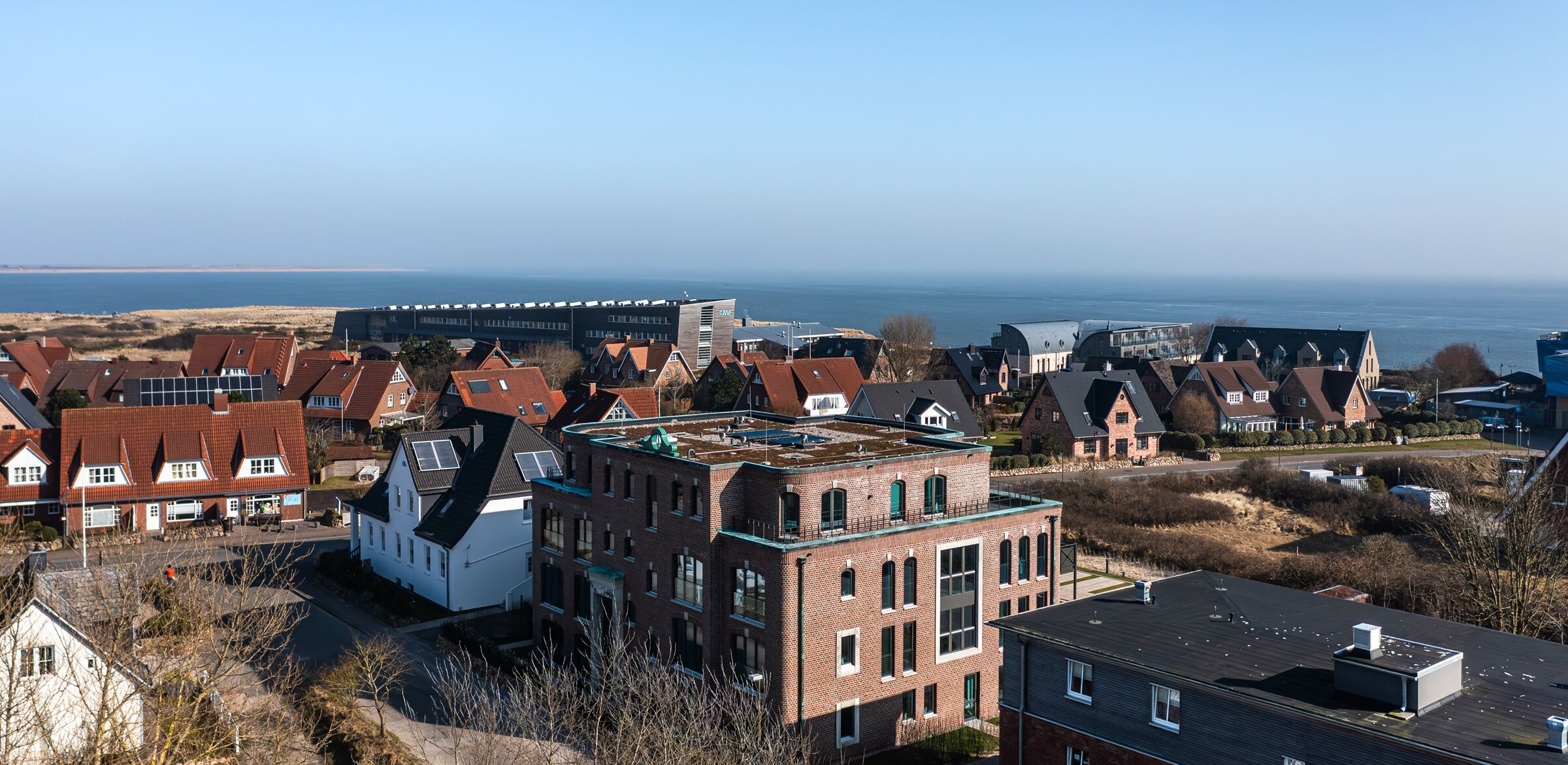 A residential area with houses, a large building with a flat roof, and a view of the sea.
