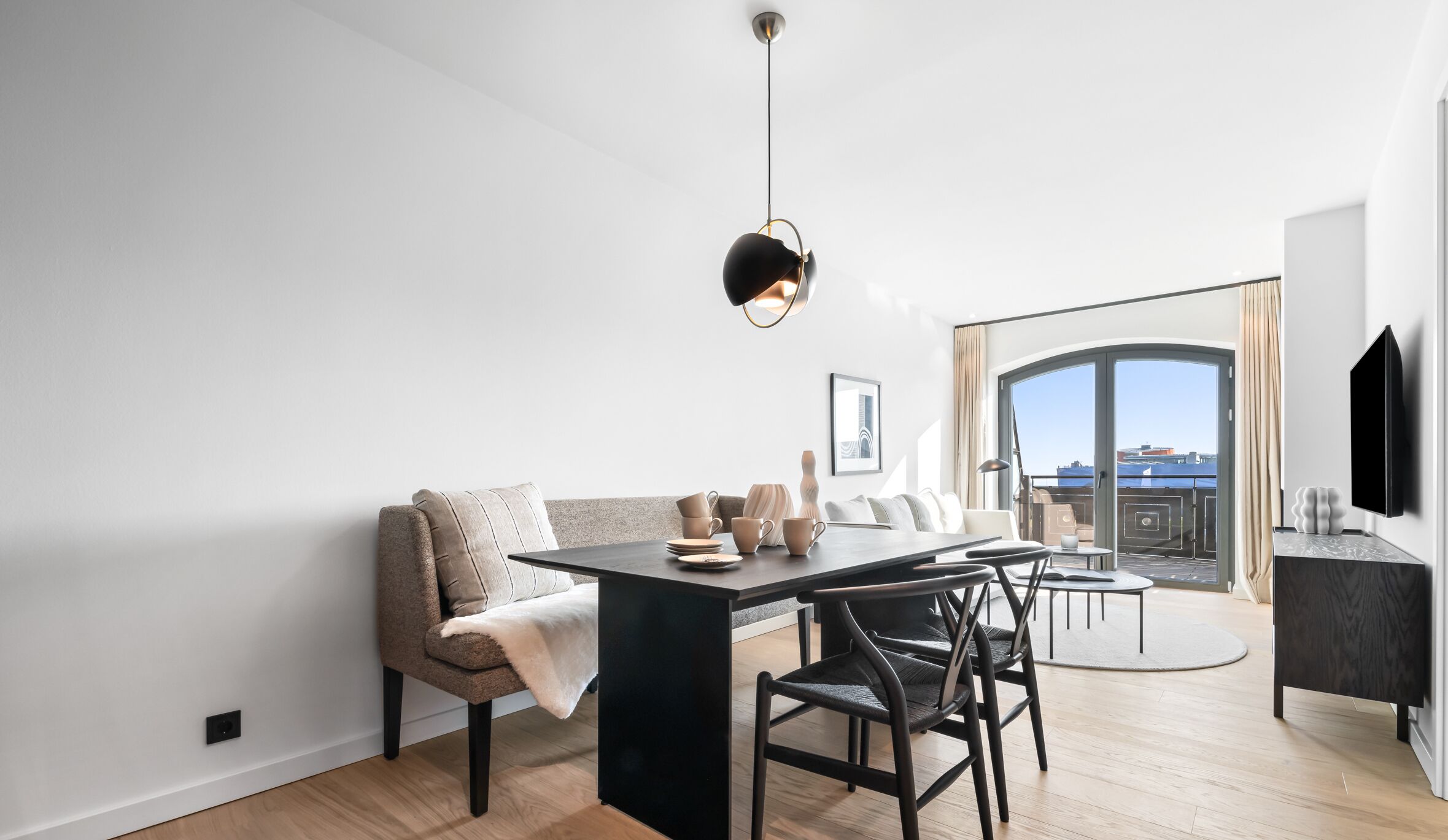 Ein moderner Essbereich mit einem schwarzen Tisch, Stühlen und einer besonderen Pendelleuchte. Eine Sitzecke und ein Balkon mit Blick sind ebenfalls zu sehen.