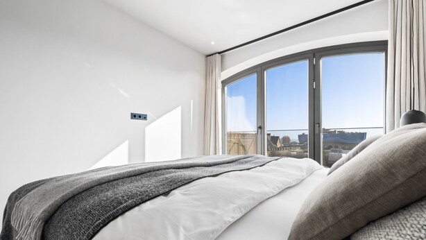 Une chambre moderne dotée d’une grande fenêtre incurvée, d’un lit agrémenté de nombreux coussins et offrant une vue sur le paysage urbain.
