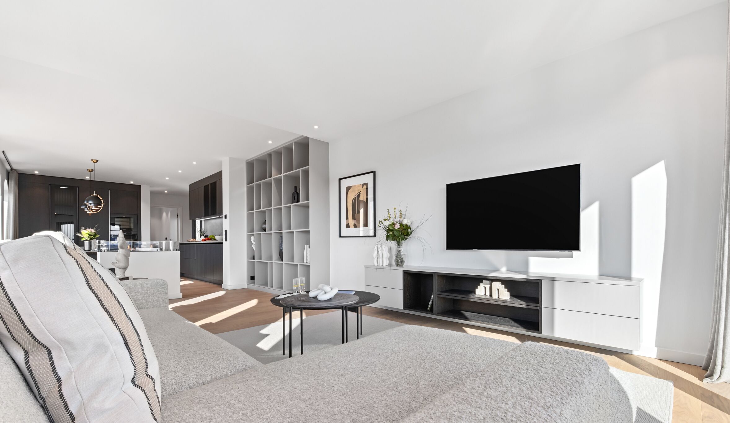 A modern living room with a large grey sofa, a flat-screen TV on a white stand, and a kitchen in the background.