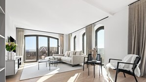 Un saló modern amb un sofà, una taula de centre i cadires, finestres grans amb cortines i vista a un edifici.