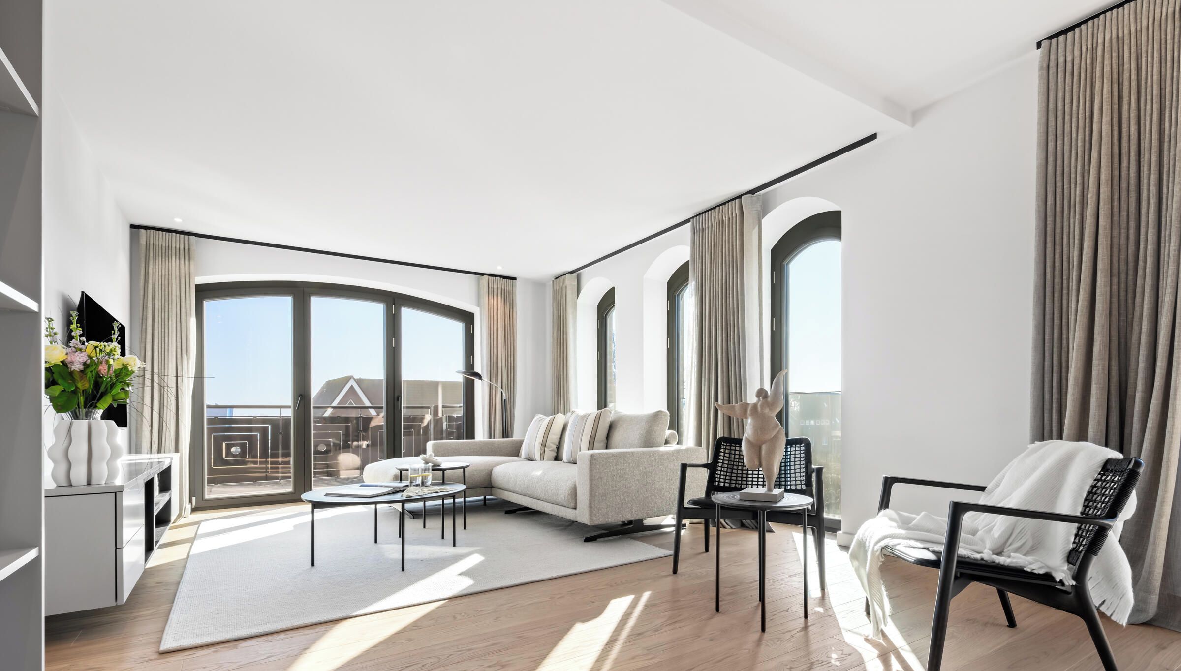 Ein modernes Wohnzimmer mit Sofa, Couchtisch und Sesseln, großen Fenstern mit Vorhängen und Blick ins Freie.