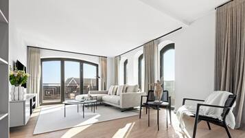 Ein modernes Wohnzimmer mit Sofa, Couchtisch und Sesseln, großen Fenstern mit Vorhängen und Blick ins Freie.