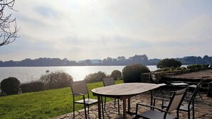 Ein Außenbereich mit Tisch und Stühlen, mit Blick auf ein Gewässer und eine entfernte Baumreihe.