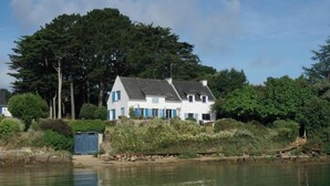 Ein weißes Haus mit grauem Dach, gelegen am Wasser, umgeben von Bäumen und Grün.