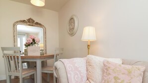 Um quarto com uma mesa de jantar, cadeiras, um sofá com almofadas florais, um espelho, um relógio de parede e um abajur.