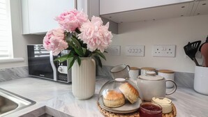 Uma cozinha com bancada de mármore, um rádio Kenwood, um vaso com flores cor-de-rosa e uma mesa posta para o café da manhã com scones e geleia.