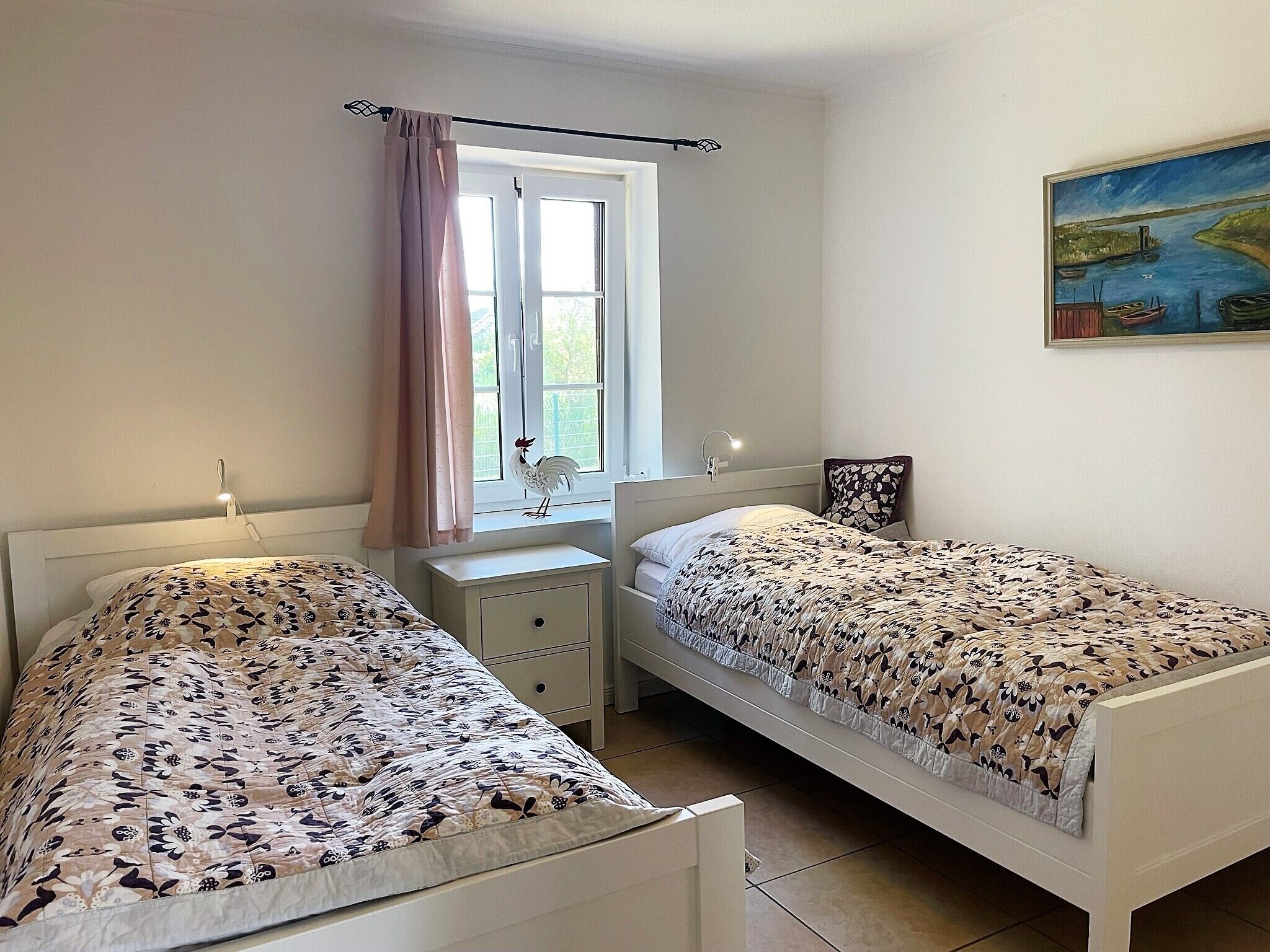 Une chambre avec deux lits simples, une fenêtre avec des rideaux, une table de chevet avec une lampe et un tableau au mur.