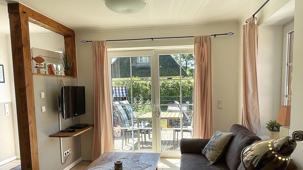 A living room with a sofa, a coffee table, a television, and a sliding glass door leading to an outdoor area with chairs and a table. - Your Perfect Getaway: Cozy Holiday Home in the Tranquil Countryside! (Rodenäs)