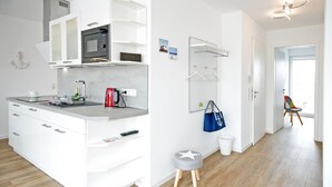 A modern kitchen with white cabinetry, a built-in microwave, and a grey countertop. There is a small stool with a star design, a white trash bin, and a blue bag hanging on the wall. - Modern penthouse (55 sqm) open all year round for 2 to 4 people in the Staffel (Norderney)