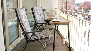 A balcony with two chairs and a table, overlooking a street with buildings and a red-brick building with a clock tower.