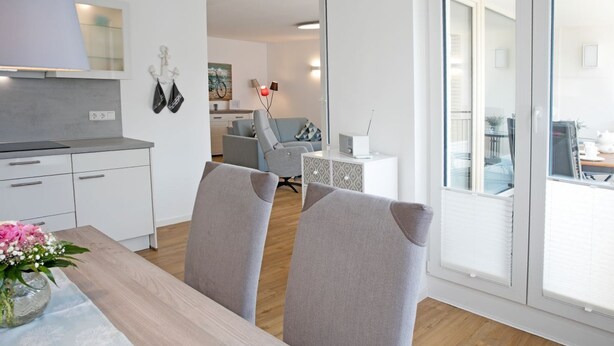 A modern kitchen with white cabinets and a wooden countertop, a dining area with grey upholstered chairs, and a view through a glass door to a balcony.