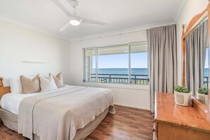 A bedroom with a large bed, a ceiling fan, a window with a view of the sea, and a wooden nightstand with a plant. - Absolute Beachfront Apartment 3T (Lennox Head)