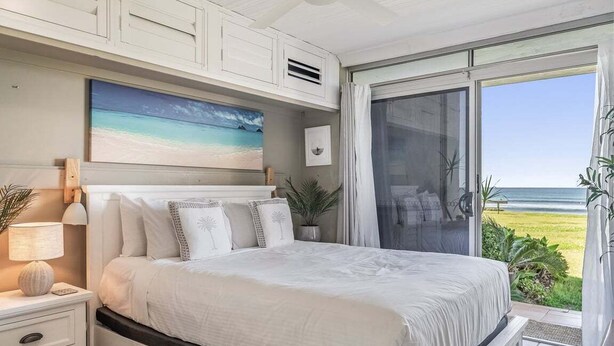 A bedroom with a large bed, a nightstand, a lamp, a painting of a beach, and a view of the ocean through a sliding door.