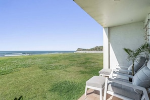 A balcony with a view of the ocean, a grassy lawn, and a seating area with cushions.