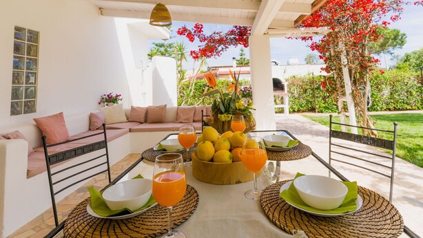 Une terrasse ensoleillée avec une table dressée pour deux personnes, agrémentée d’un panier de fruits, de verres de jus d’orange et de bols blancs posés sur des sets de table en fibres tressées.