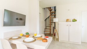 Un coin repas avec une table dressée pour quatre personnes, un téléviseur fixé au mur, un escalier et un meuble blanc agrémenté d’objets décoratifs.