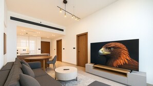 Ein modernes Wohnzimmer mit großem Flachbildfernseher, einer Couch, einem Couchtisch und einem Essbereich mit Küche im Hintergrund.