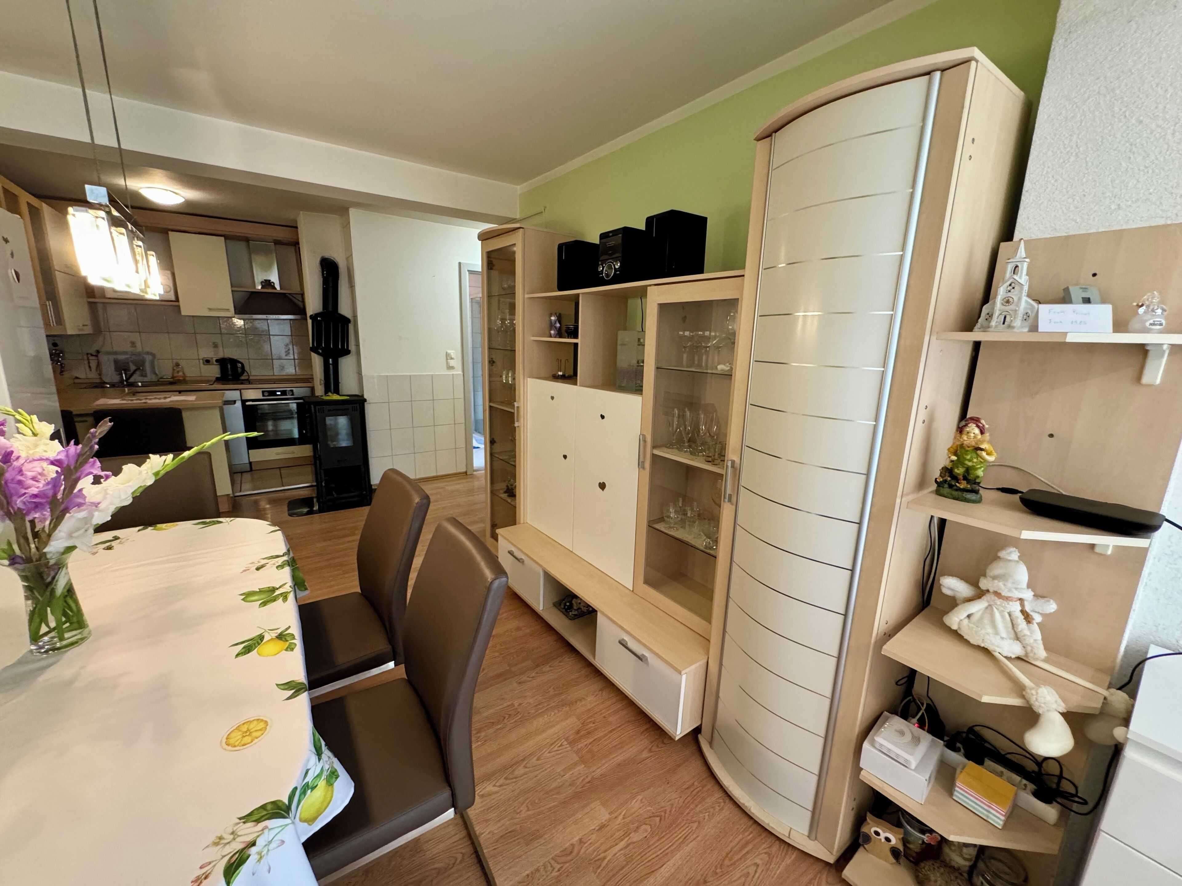 A modern kitchen with a dining area, featuring a wooden table with a floral tablecloth, a built-in cabinet with glass doors, and a small statue on a shelf.