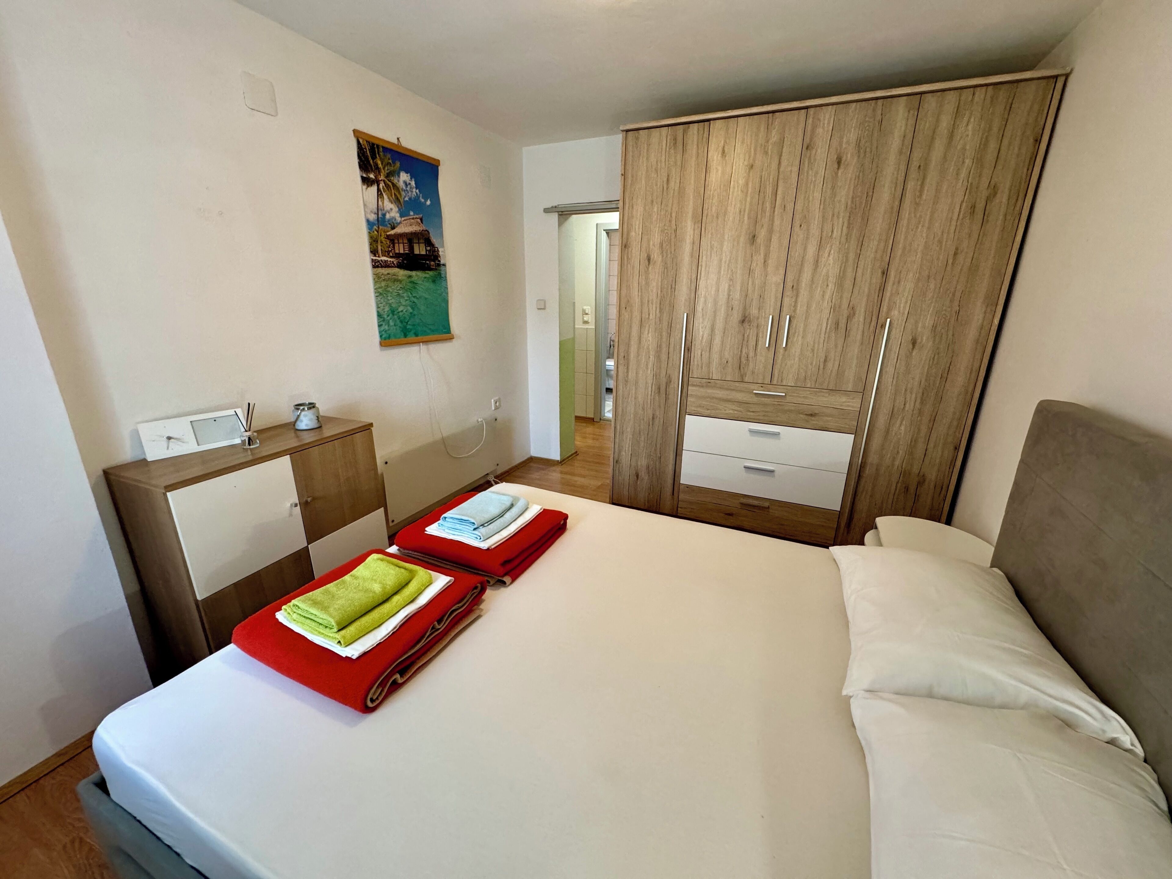 Une chambre d’hôtel avec un lit, une armoire en bois, un petit bureau avec une chaise, un tableau accroché au mur et une salle de bain visible à travers une porte ouverte.