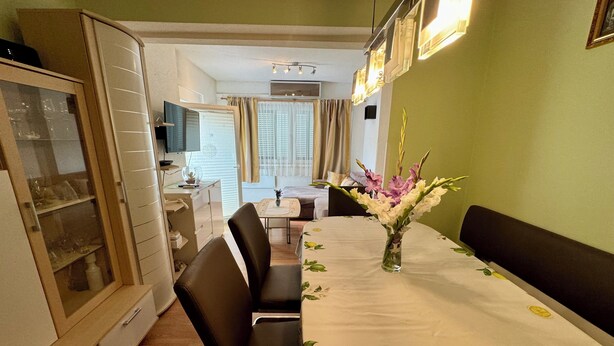 A dining area with a table set for four, a vase of flowers, and a television in the background. - Apartment Fruk 5 (Jadranovo)