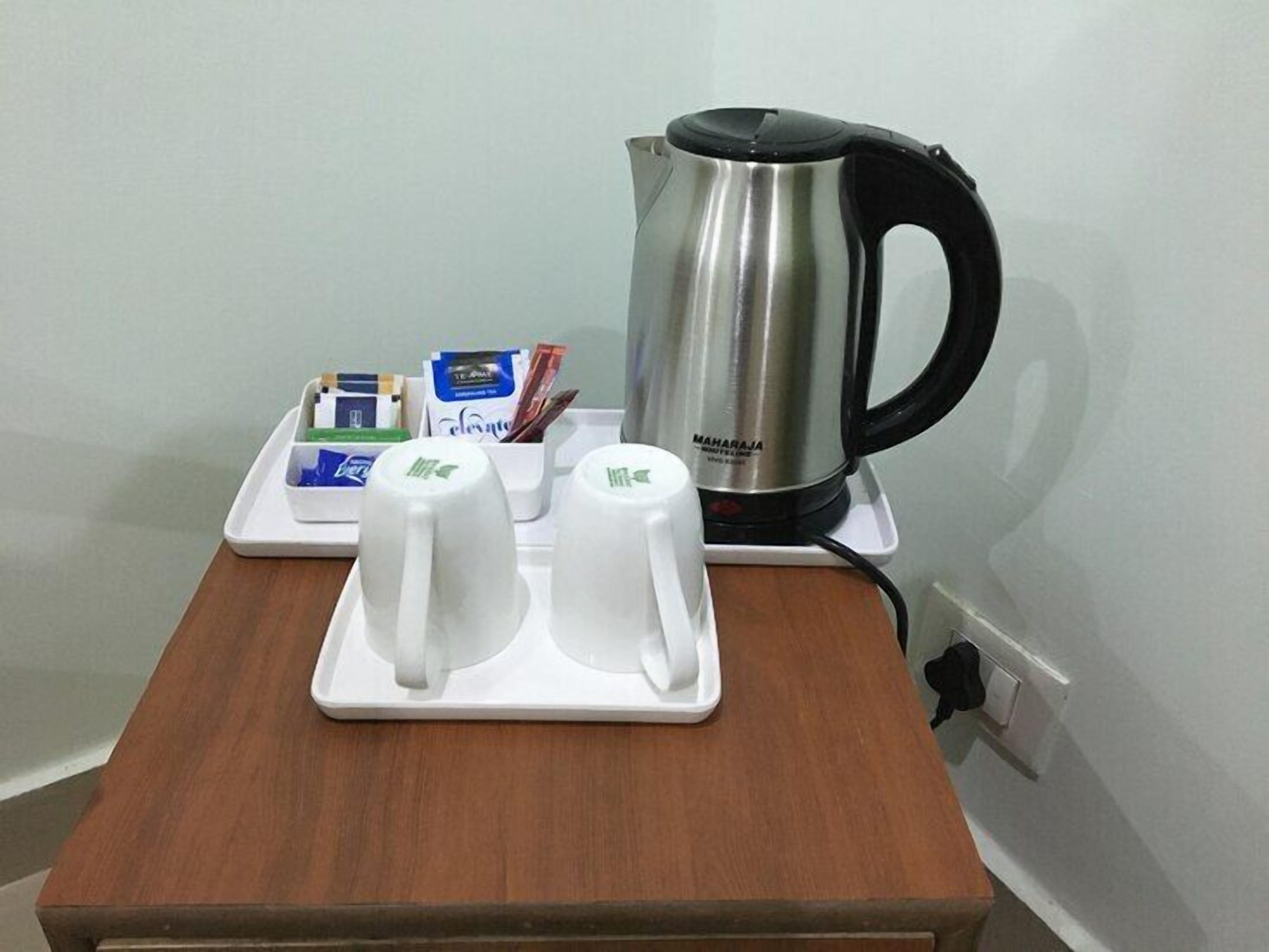 Una estación de café con una tetera de acero inoxidable, dos tazas blancas y una bandeja con bolsitas de té y una caja de pañuelos.