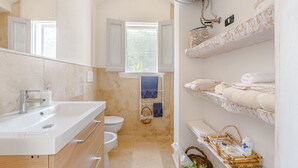 Uma casa de banho moderna com uma sanita, chuveiro e lavatório branco. Toalhas e produtos de higiene pessoal estão dispostos em prateleiras.