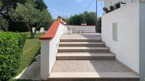 A white building with a red roof, a set of beige steps leading to a patio area with outdoor furniture, and a well-maintained garden with various plants and trees. - Agriturismo Perla del Salento, just a few steps from the sea and the beautiful beaches (Puglia)