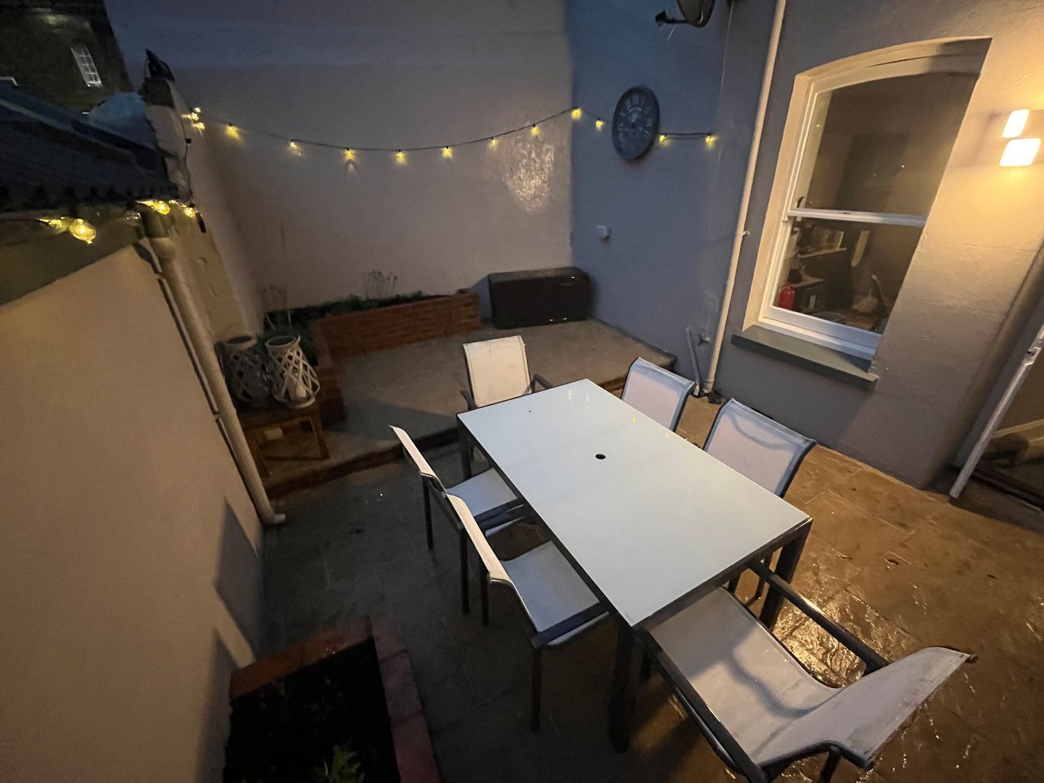 Un patio con mesa y sillas blancas, luces colgantes, un reloj y una ventana.