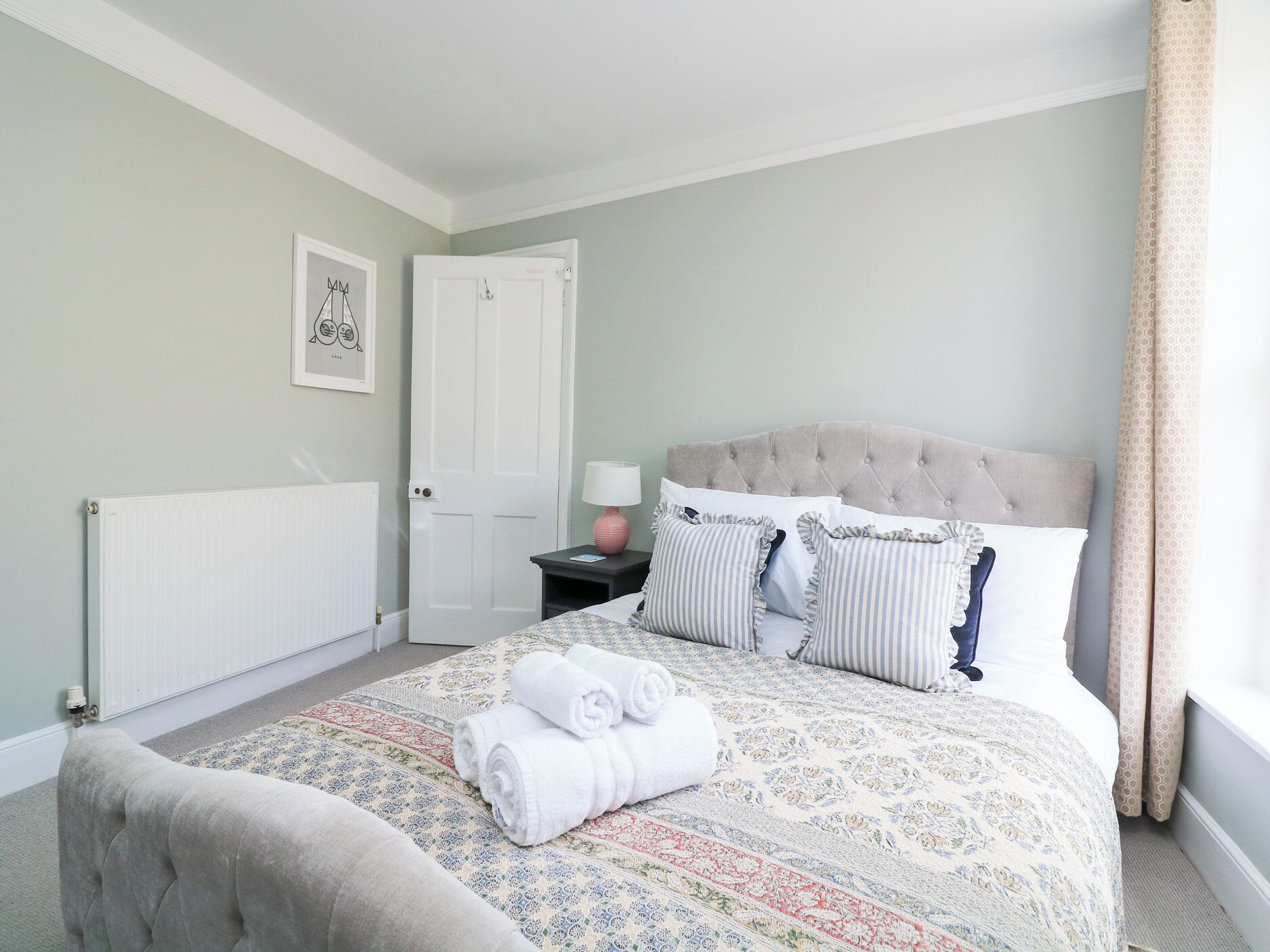 Een slaapkamer met een gebobbelde hoofdbord, een bed met een decoratieve spreideken, een nachtkastje met een lamp en een opgerolde handdoek op het bed.
